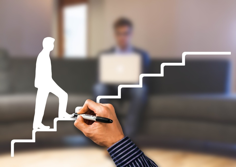Drawn white staircase with person ascending superimposed on blurred background where a person is seated and working on a laptop. Header image for "growth strategies"